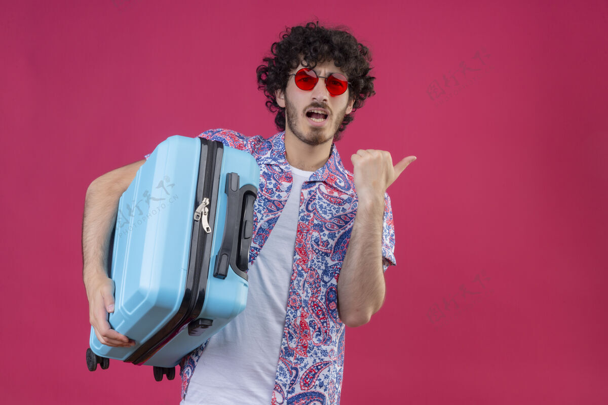手提箱印象深刻的年轻英俊的卷发旅行者男子戴着太阳镜拿着手提箱 指着右边的粉红色空间和复印空间指向太空旅行者