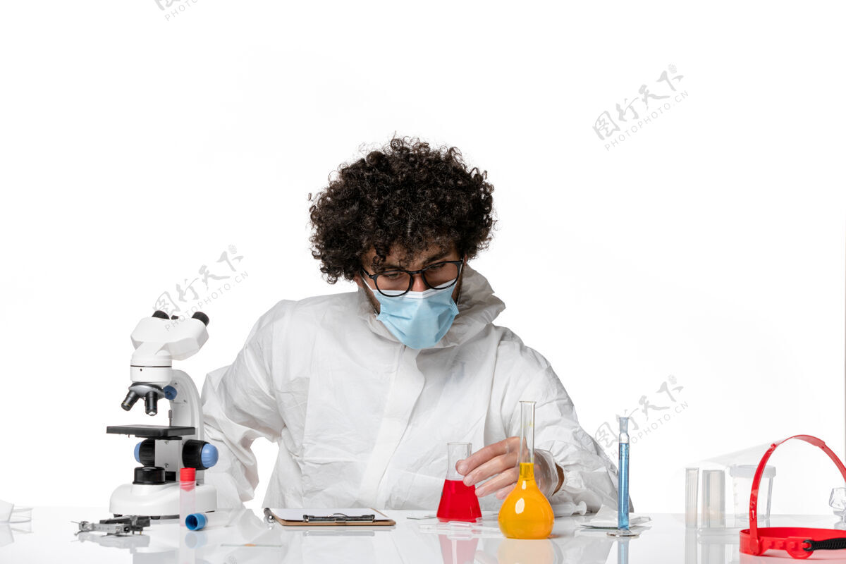保持正面图：男医生穿着防护服 戴着口罩 拿着淡白色背景上的红色溶液瓶——流行性冠状病毒浅白色实验室外套视图