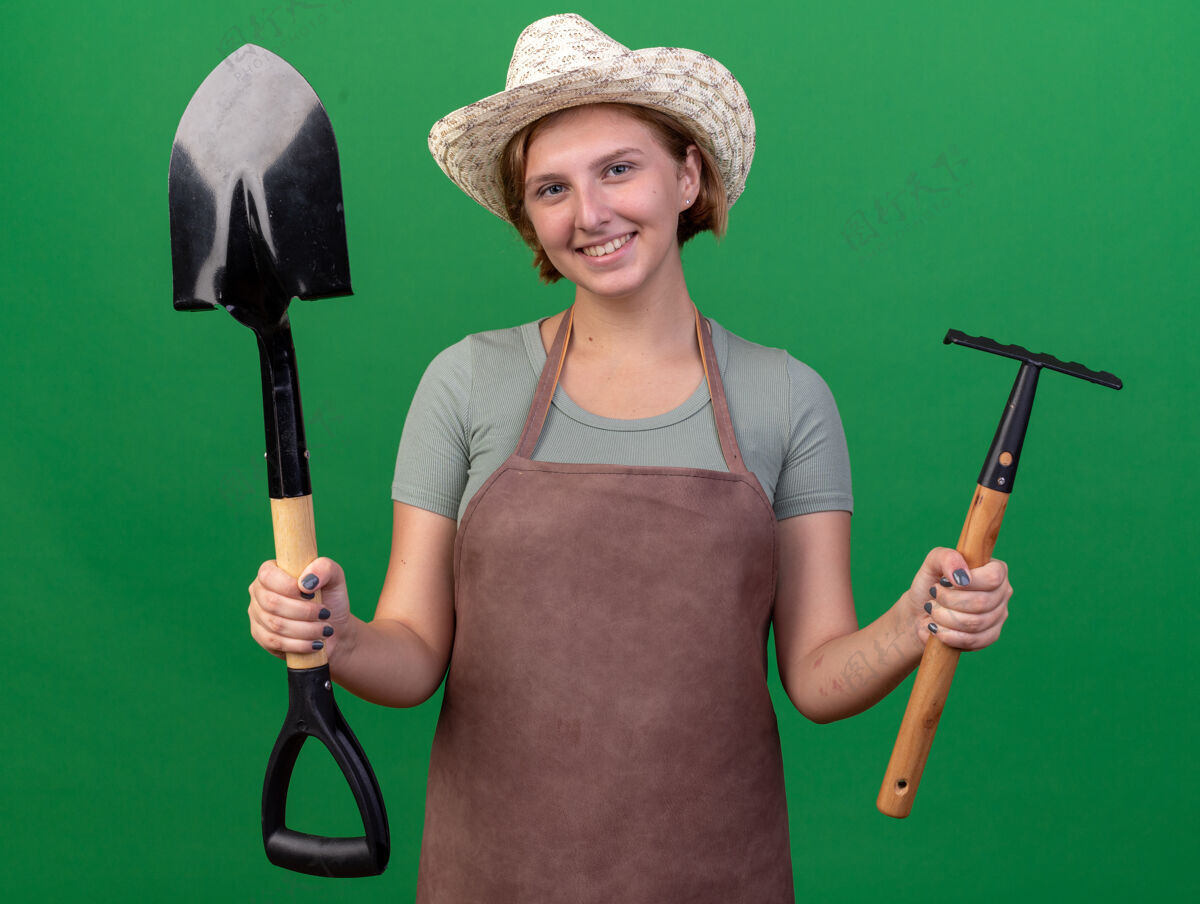 年轻的微笑着的年轻的斯拉夫女园丁戴着园艺帽拿着铲子和耙子在草地上帽子女的园艺