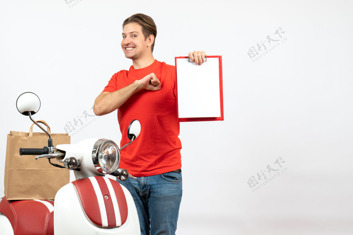 健身身穿红色制服的年轻自豪的送货员站在滑板车旁 在白墙上展示文件人年轻站