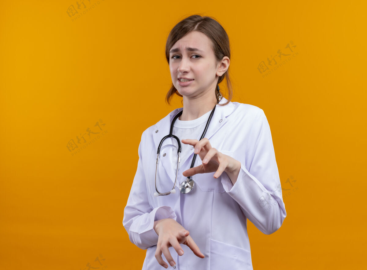 长袍恼怒的年轻女医生穿着医用长袍和听诊器在孤立的橙色空间与复印空间太空听诊器橙色