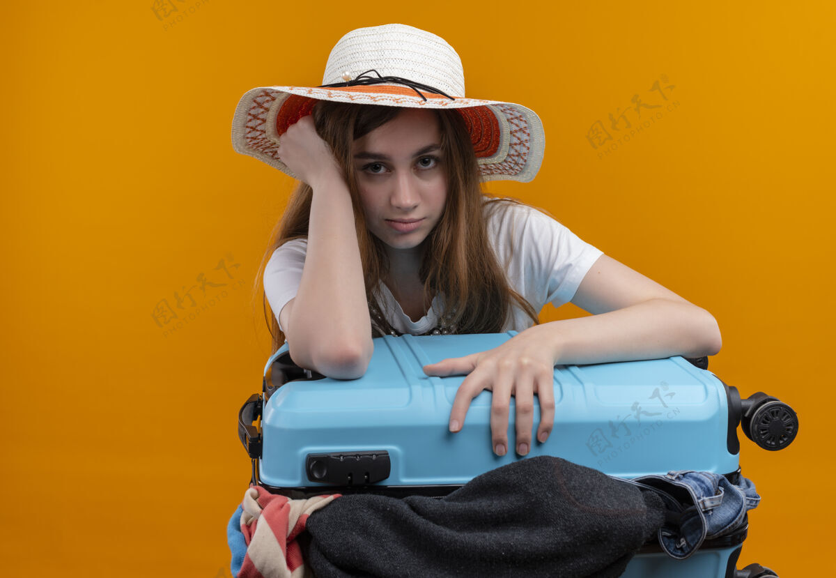 手无聊的年轻旅行家女孩戴着帽子 提着装满衣服的手提箱 手放在头上 在孤立的橙色空间和复制空间衣服无聊复制