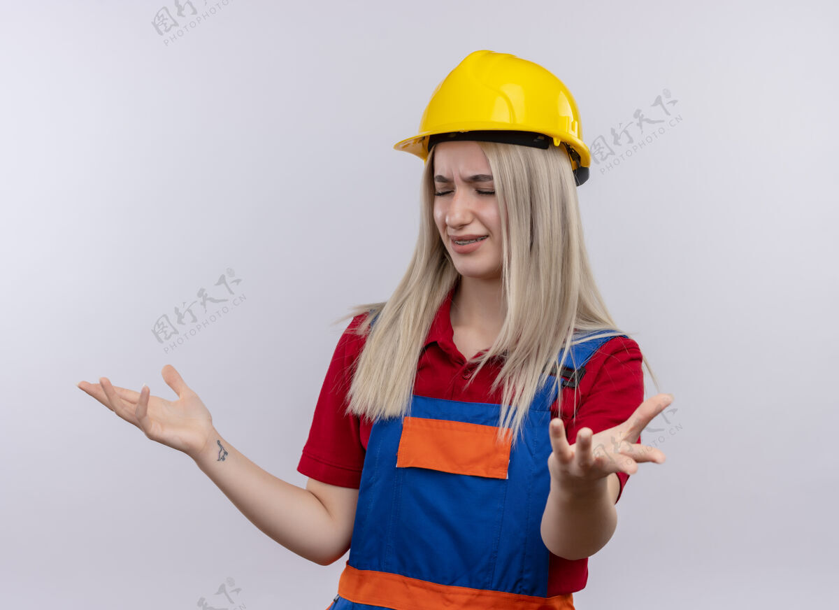 年轻恼怒的年轻金发工程师建筑工女孩穿着制服 戴着牙套 在孤立的空白处空手而归工程师金发制服