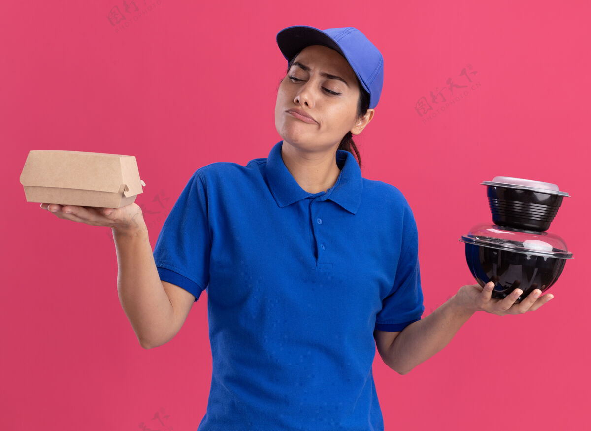 帽子想着那个穿着制服 戴着帽子的年轻送货女孩 看着隔离在粉红色墙上的食品容器感觉表情递送