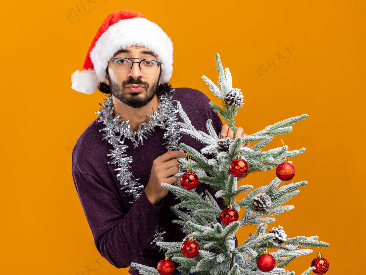 脖子快乐的年轻帅哥站在圣诞树后面 戴着圣诞帽 脖子上戴着花环 橙色的背景孤立着小伙子圣诞快乐圣诞树