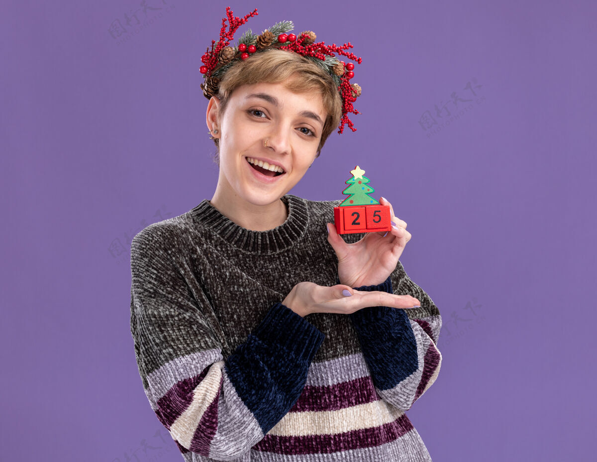 头快乐的年轻漂亮女孩戴着圣诞花环拿着圣诞树玩具和日期指着它看着隔离在紫色背景上的相机指向举行日期