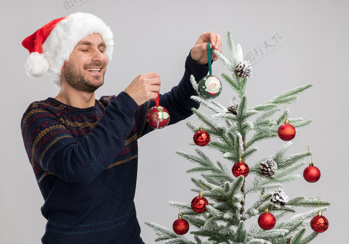 快乐快乐的白人年轻人戴着圣诞帽站在圣诞树旁 用圣诞装饰球装饰圣诞树 在白色背景上闭着眼睛大笑男人圣诞封闭