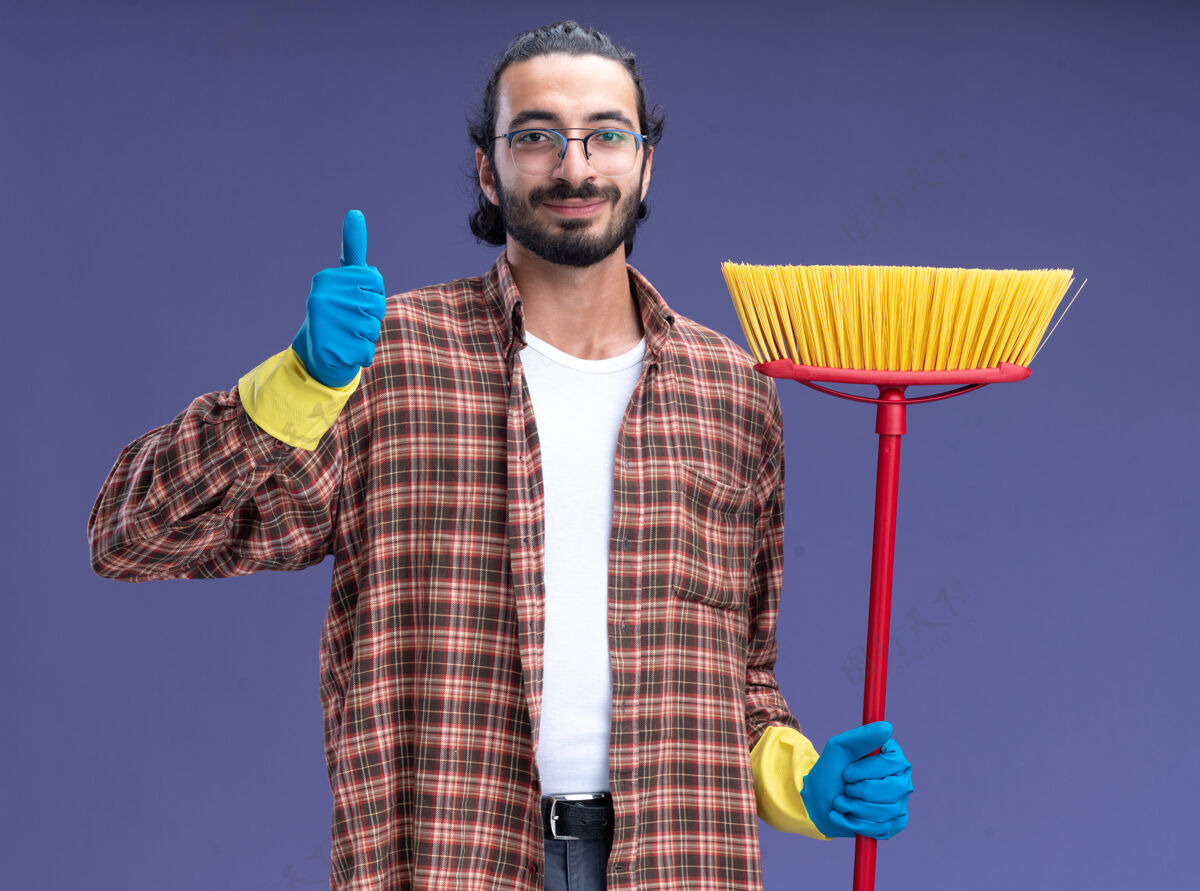 穿高兴的年轻帅气的清洁工穿着t恤 戴着手套 拿着拖把 大拇指向上伸出 孤零零地站在蓝色的墙上脸人男人