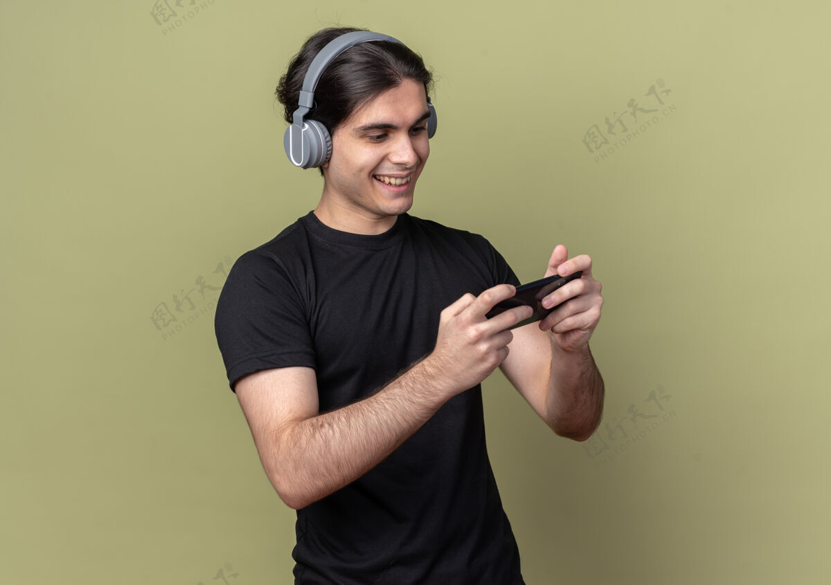 脸带着微笑的年轻帅哥穿着黑色t恤戴着耳机在橄榄绿的墙上玩游戏姿势站着微笑