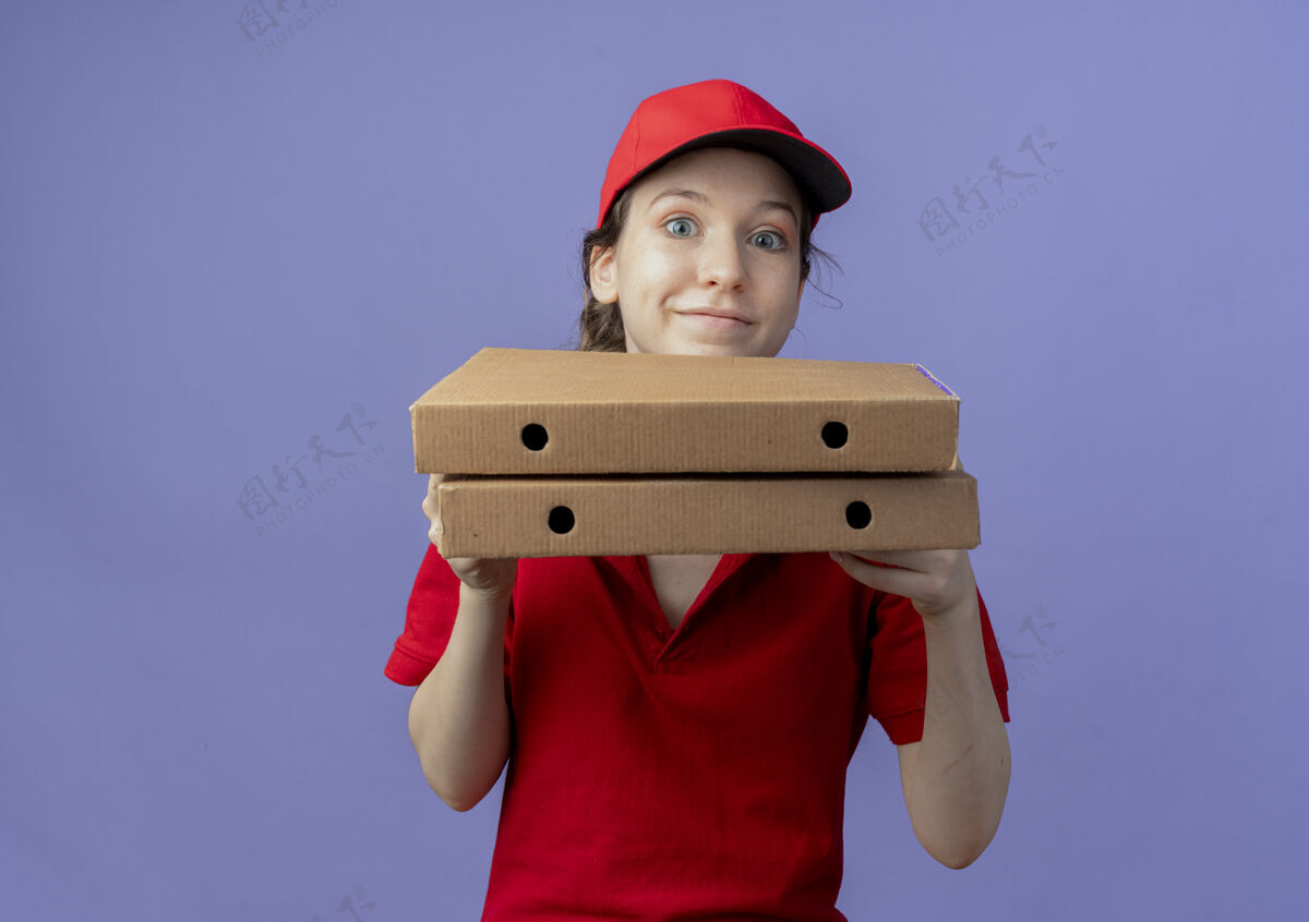 紫色很高兴年轻漂亮的送货女孩穿着红色制服 戴着帽子拿着披萨包 看着隔离在紫色背景上的相机 还有复印空间帽子女孩背景
