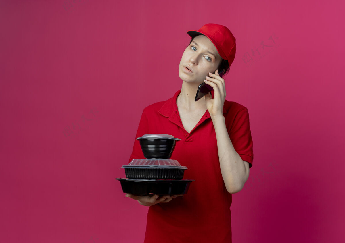 帽子令人印象深刻的年轻漂亮的送货女孩穿着红色制服 戴着帽子讲电话 拿着食品容器 看着摄像机在深红的背景下与复制空间隔离电话手持年轻