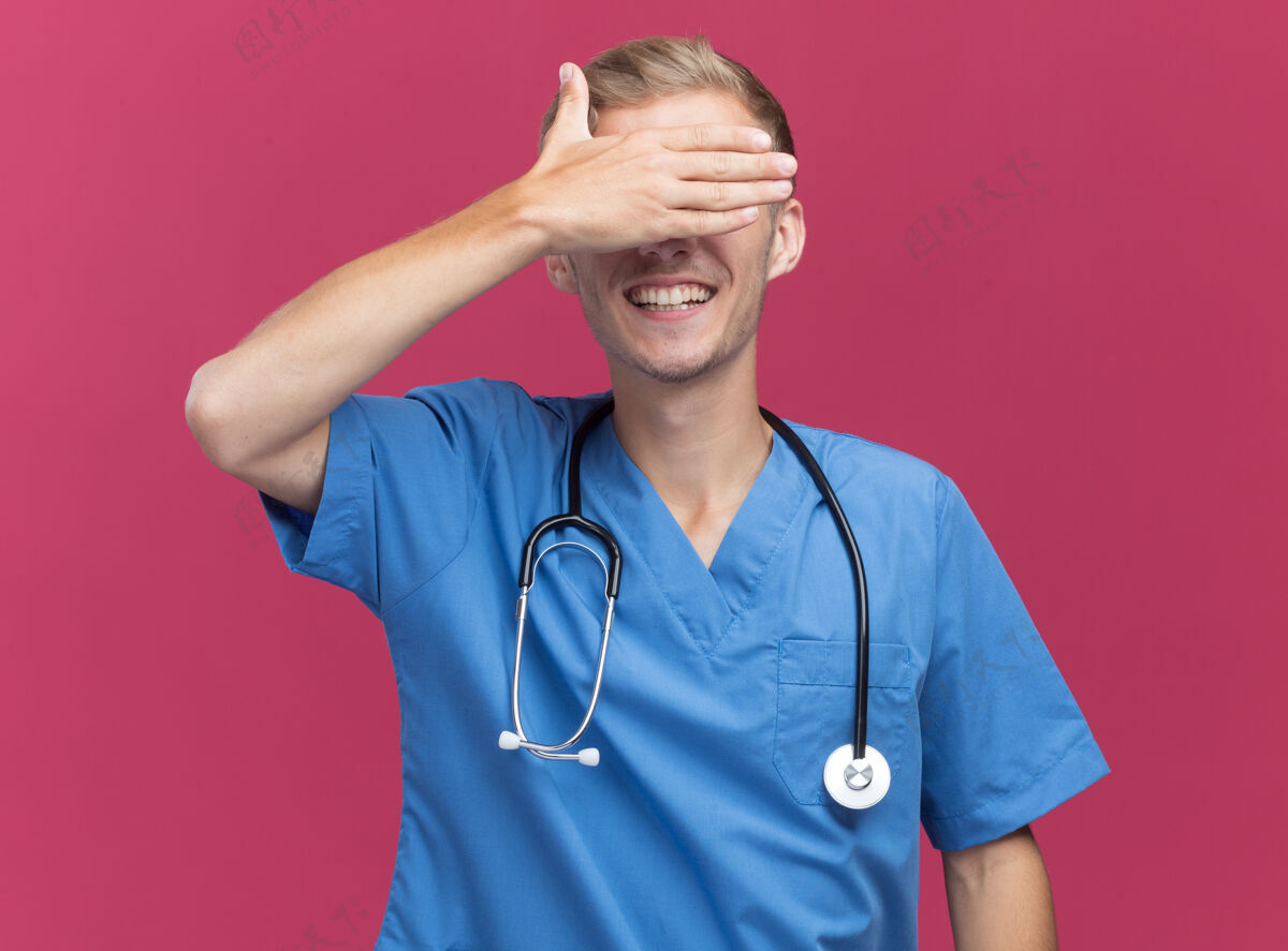 人面带微笑的年轻男医生 穿着医生制服 戴着听诊器 眼睛蒙着 手被隔离在粉红色的墙上衣服掩护站着