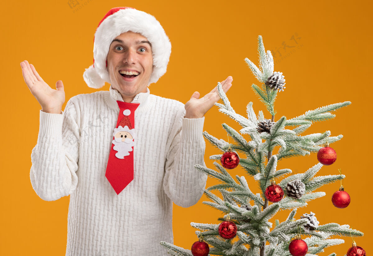 年轻人年轻帅哥戴着圣诞帽 打着圣诞老人的领带 站在装饰好的圣诞树旁 看着镜头 两手空空的 孤立在橙色的背景上小伙子空的圣诞快乐