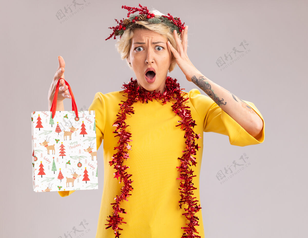 圣诞快乐关心的金发女郎头戴圣诞花环 脖子上戴着金属丝花环 手里拿着圣诞礼品袋 看着相机 抚摸着隔离在白色背景上的头举行白色周围