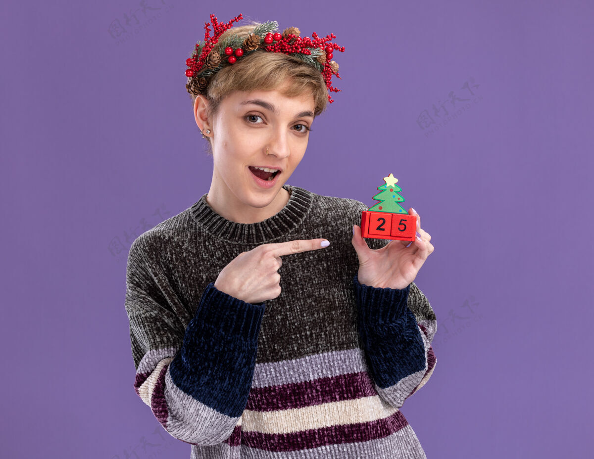 年轻给人印象深刻的年轻漂亮女孩戴着圣诞花环拿着圣诞树玩具和日期指着它看着隔离在紫色背景上的相机头花圈日期