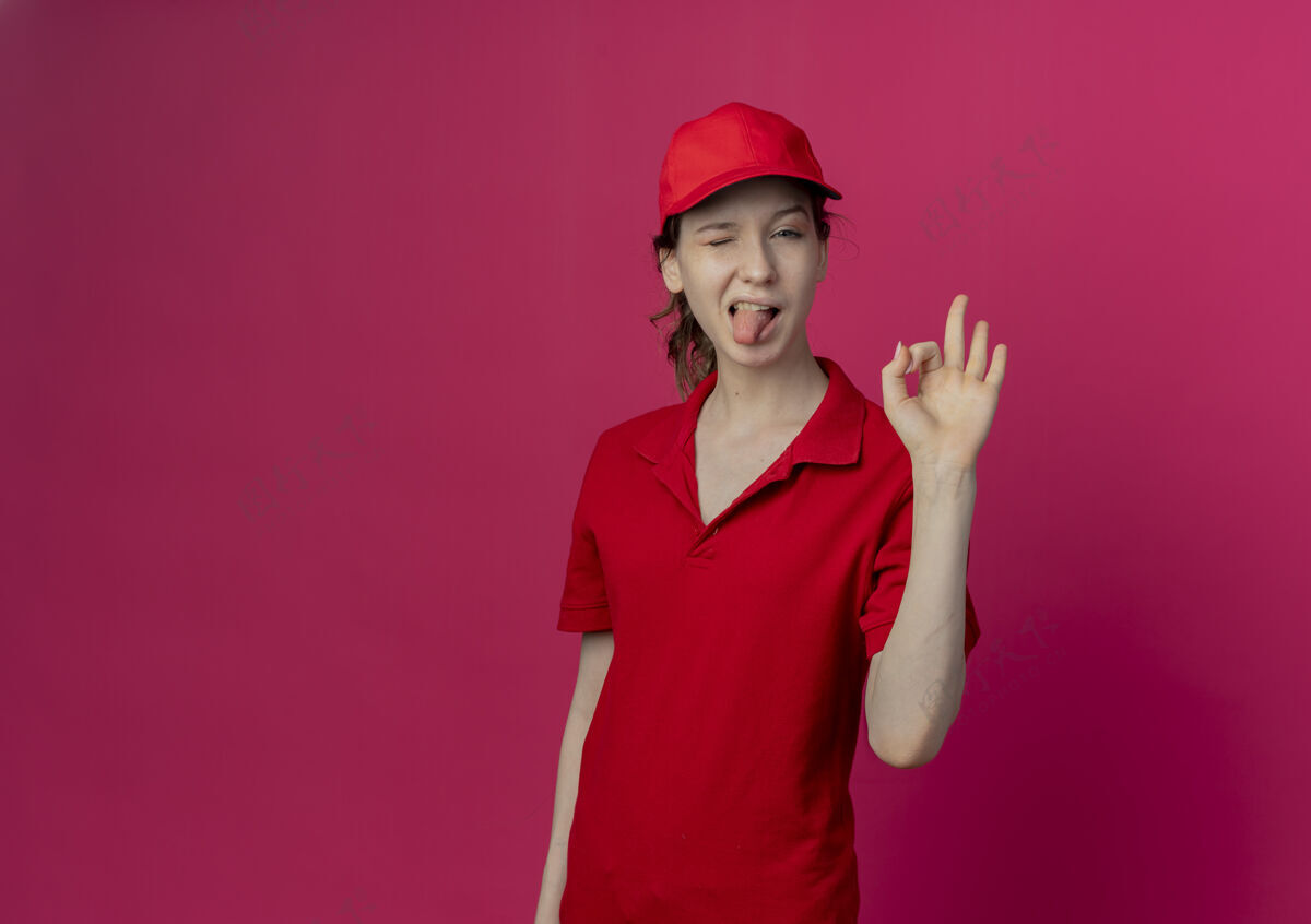 标志自信的年轻漂亮的送货女孩 穿着红色制服 戴着帽子 做着“ok”的手势 眨着眼睛 在深红色的背景上露出孤立的舌头 还有复制空间女孩红色深红色