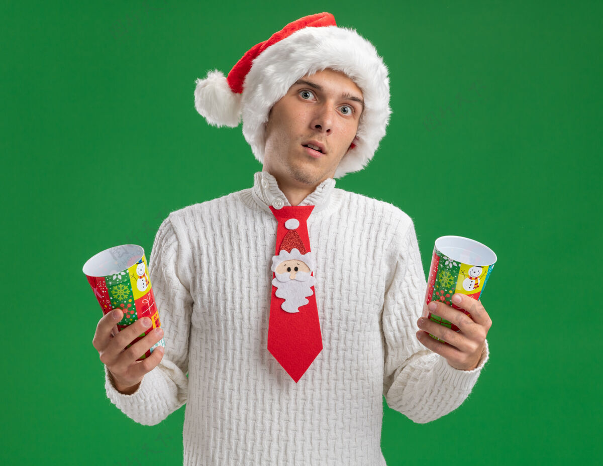 领带印象深刻的年轻帅哥戴着圣诞帽打着圣诞老人的领带拿着塑料圣诞杯看着隔离在绿色背景上的相机绿色塑料年轻