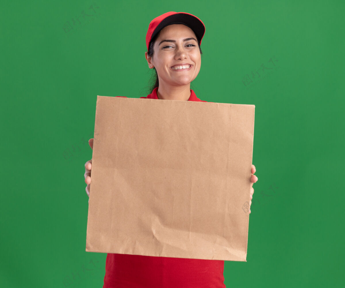 微笑面带微笑的年轻送货女孩穿着制服 戴着帽子 在绿色的墙壁上孤立地站在前面 手里拿着纸制食品包抱着人包装