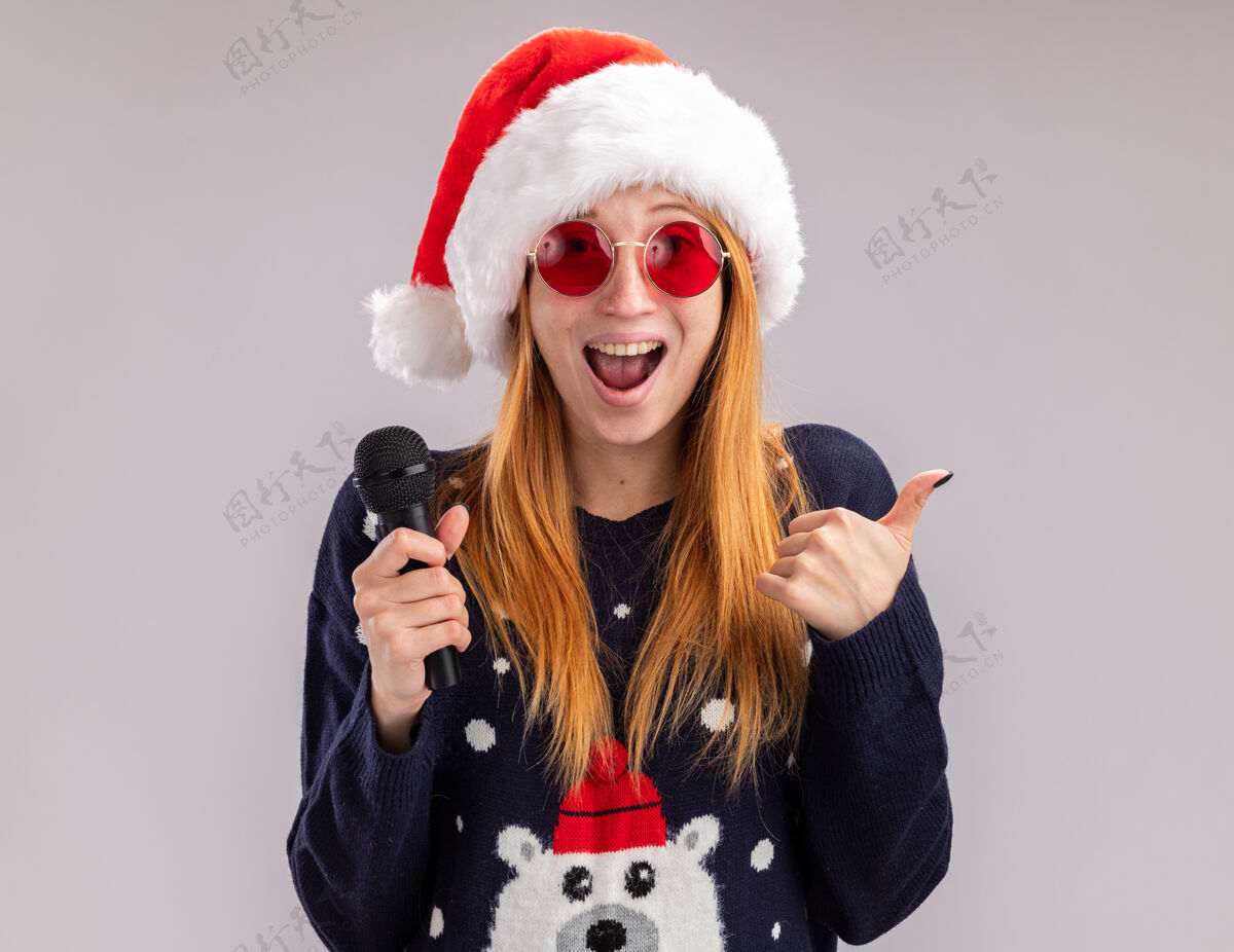 帽子兴奋的年轻漂亮女孩戴着圣诞帽戴着眼镜拿着麦克风在白色背景上孤立地竖起大拇指圣诞快乐显示年轻