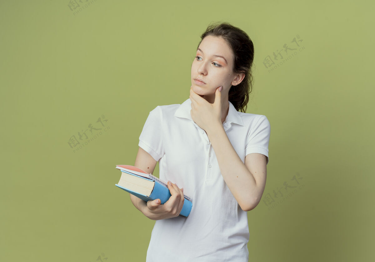 学生体贴的年轻漂亮的女学生拿着书和记事本 看着侧面 触摸着橄榄绿背景上孤立的脸 还有复印空间触摸笔记背景
