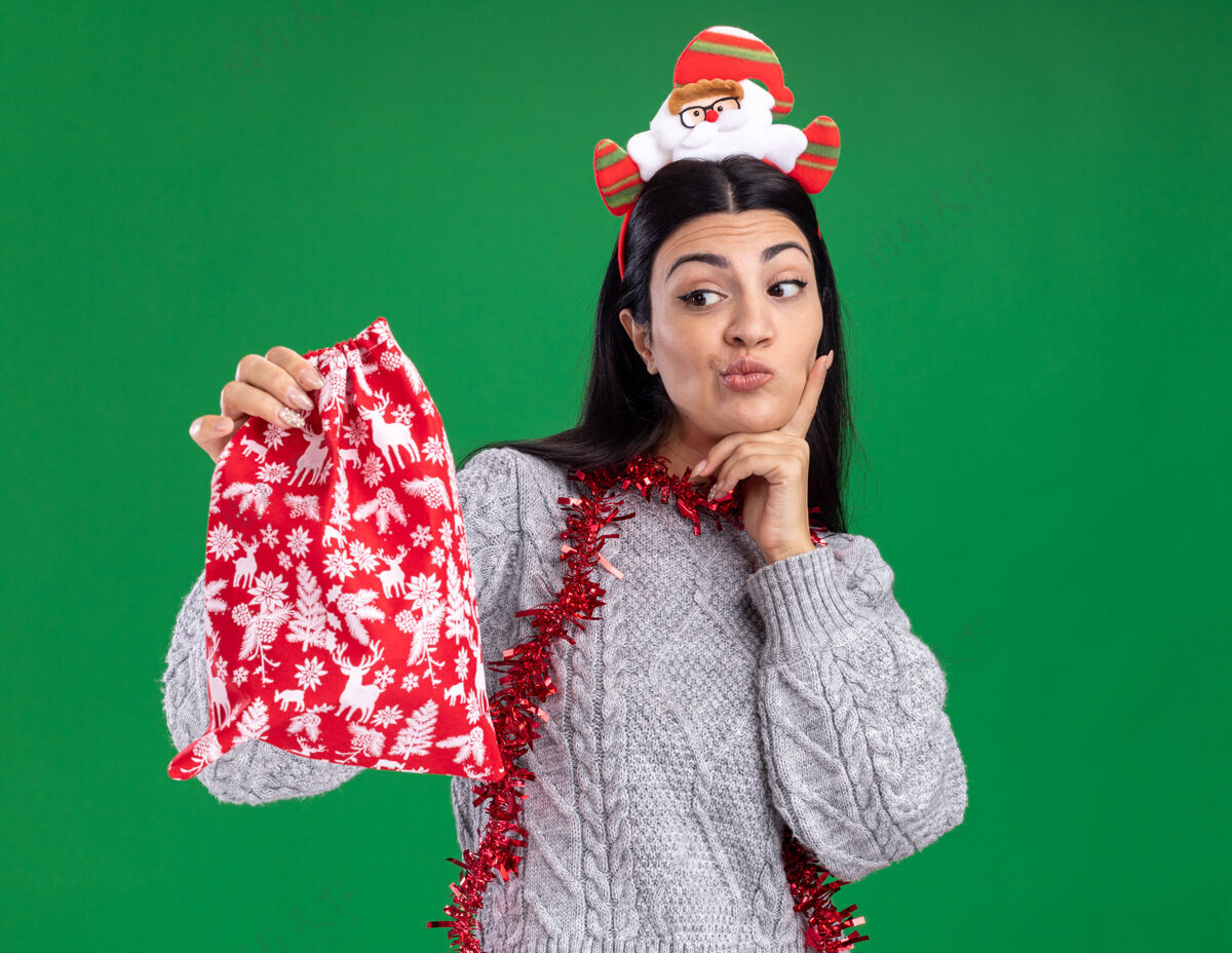 袋子可疑的年轻白人女孩戴着圣诞老人的头带 脖子上戴着金箔花环 手放在下巴上 手放在绿色背景上 看着圣诞礼物袋头带圣诞快乐圣诞老人