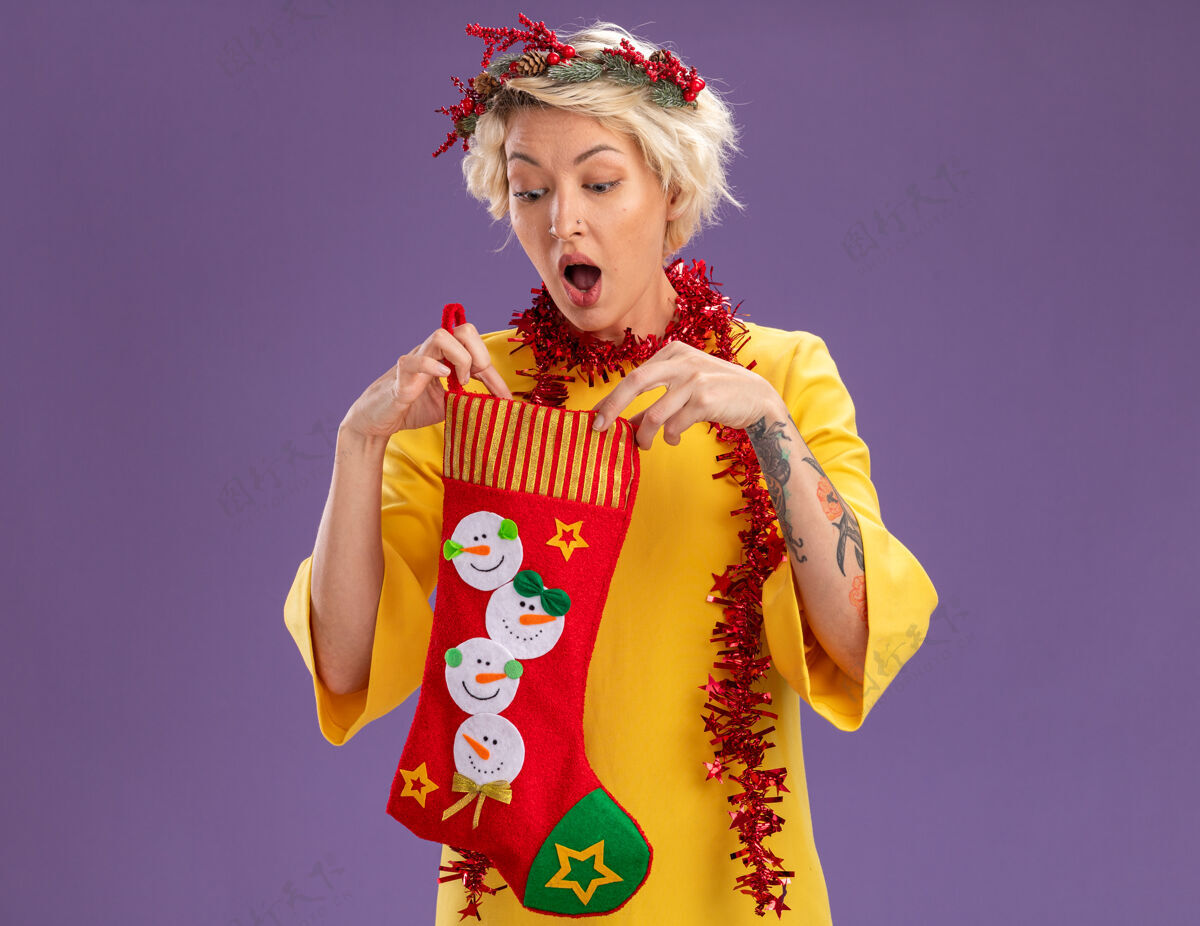 金属丝好奇的金发女郎头戴圣诞花环 脖子上戴着金箔花环 手里拿着圣诞长袜 在紫色的背景下孤立地看着里面好奇头圣诞快乐