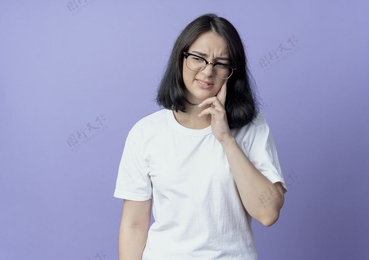 手指年轻漂亮的白人女孩戴着眼镜 手指放在脸颊上 看着牙痛的一边 紫色背景上有空白背景痛苦漂亮