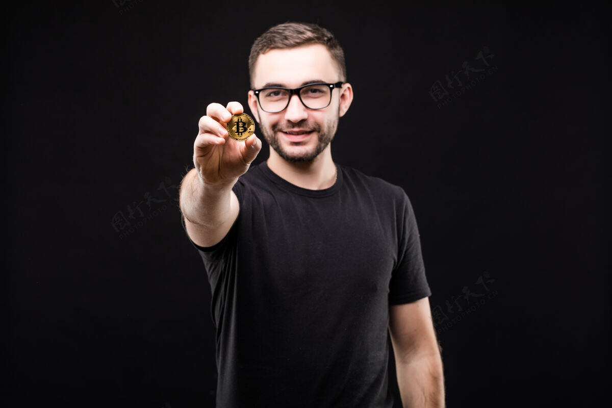 黑暗照片中的年轻帅哥戴着眼镜 穿着黑色衬衫 在相机上用金色比特币指着黑色手比特币经济