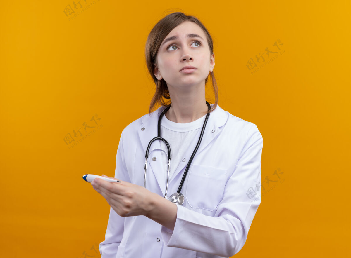 医生焦虑的年轻女医生穿着医用长袍和听诊器拿着温度计在孤立的橙色空间穿橙色女