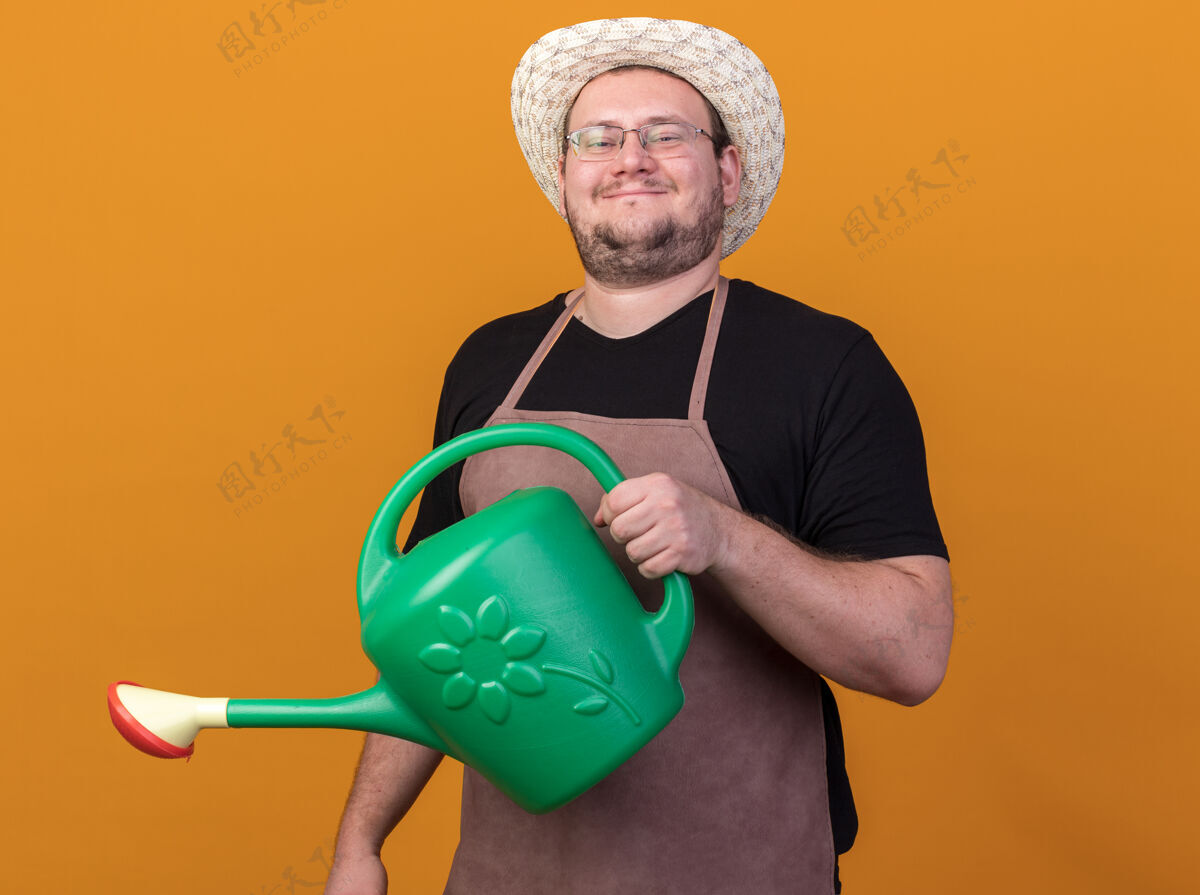 姿势高兴的年轻男园丁戴着园艺帽拿着浇水罐隔离在橙色的墙上请浇水男性
