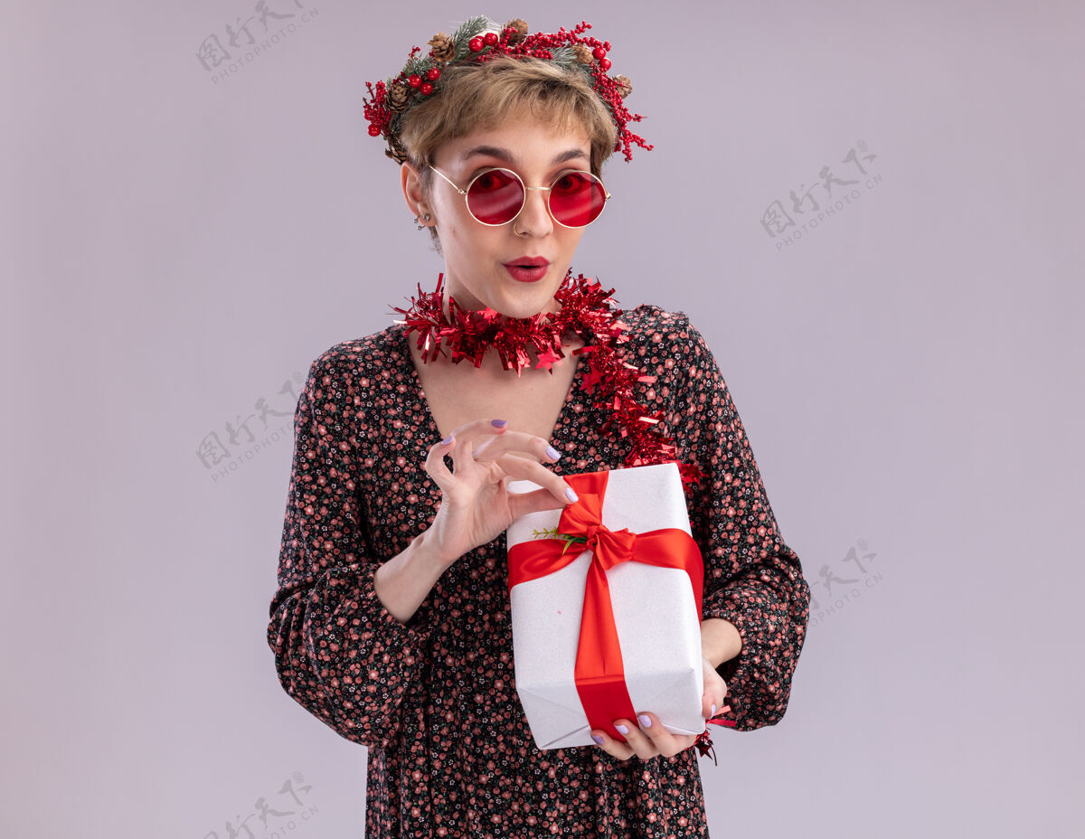 好奇好奇的年轻漂亮女孩戴着圣诞花环 脖子上戴着金箔花环 戴着眼镜 手里拿着礼包 手里拿着丝带 看着隔离在白色背景上的相机圣诞快乐空间头