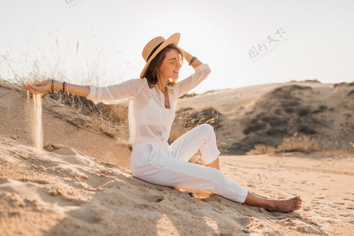 度假落日时分 一个穿着白色衣服 戴着草帽和太阳镜 在沙漠沙滩上摆造型的时髦迷人的微笑女人波西米亚沙漠衬衫