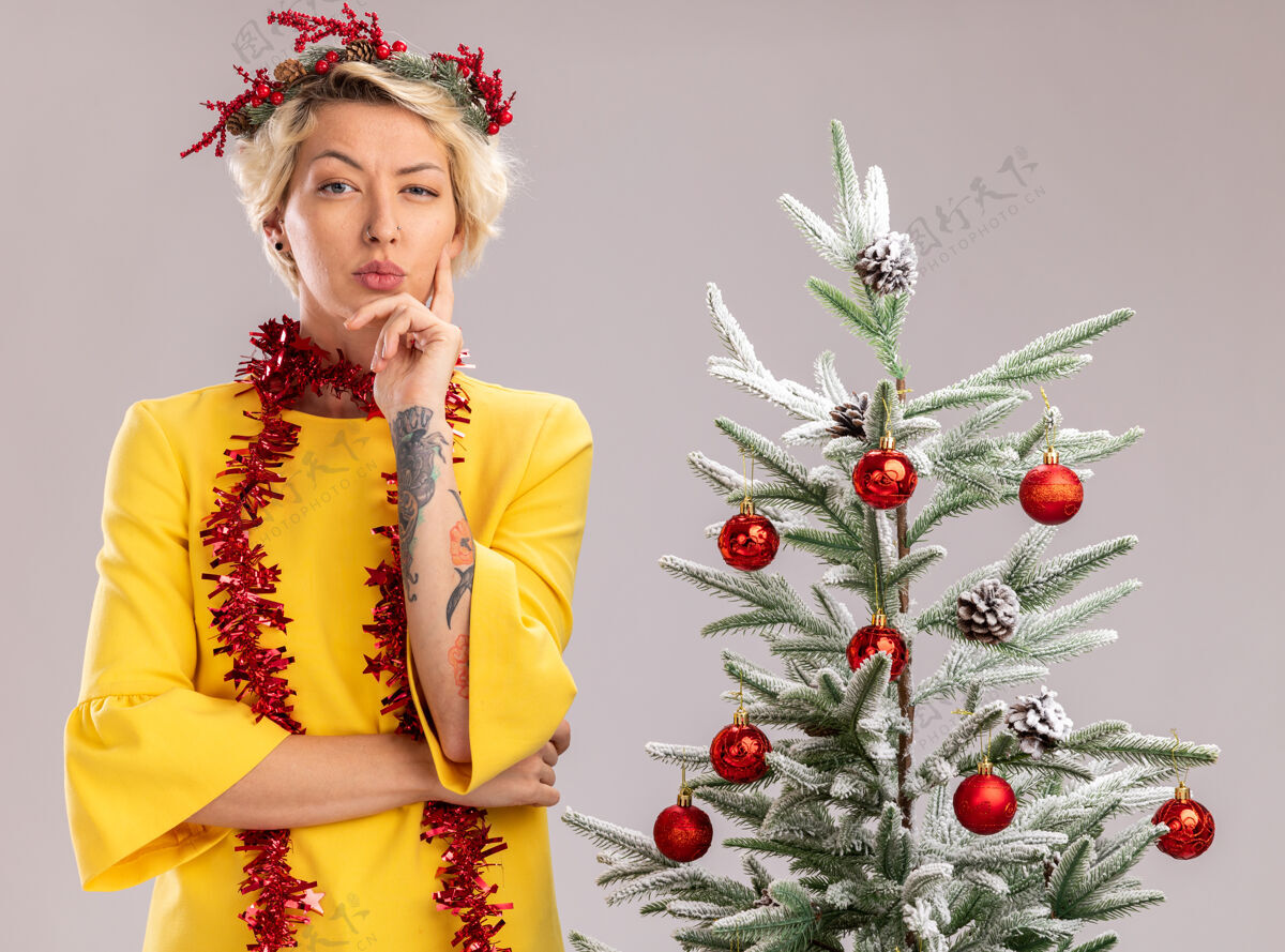 圣诞树戴着圣诞花环 脖子上戴着金箔花环的可疑金发女郎站在装饰好的圣诞树旁 看着相机 手放在下巴上 在白色背景下保持隔离站着圣诞怀疑
