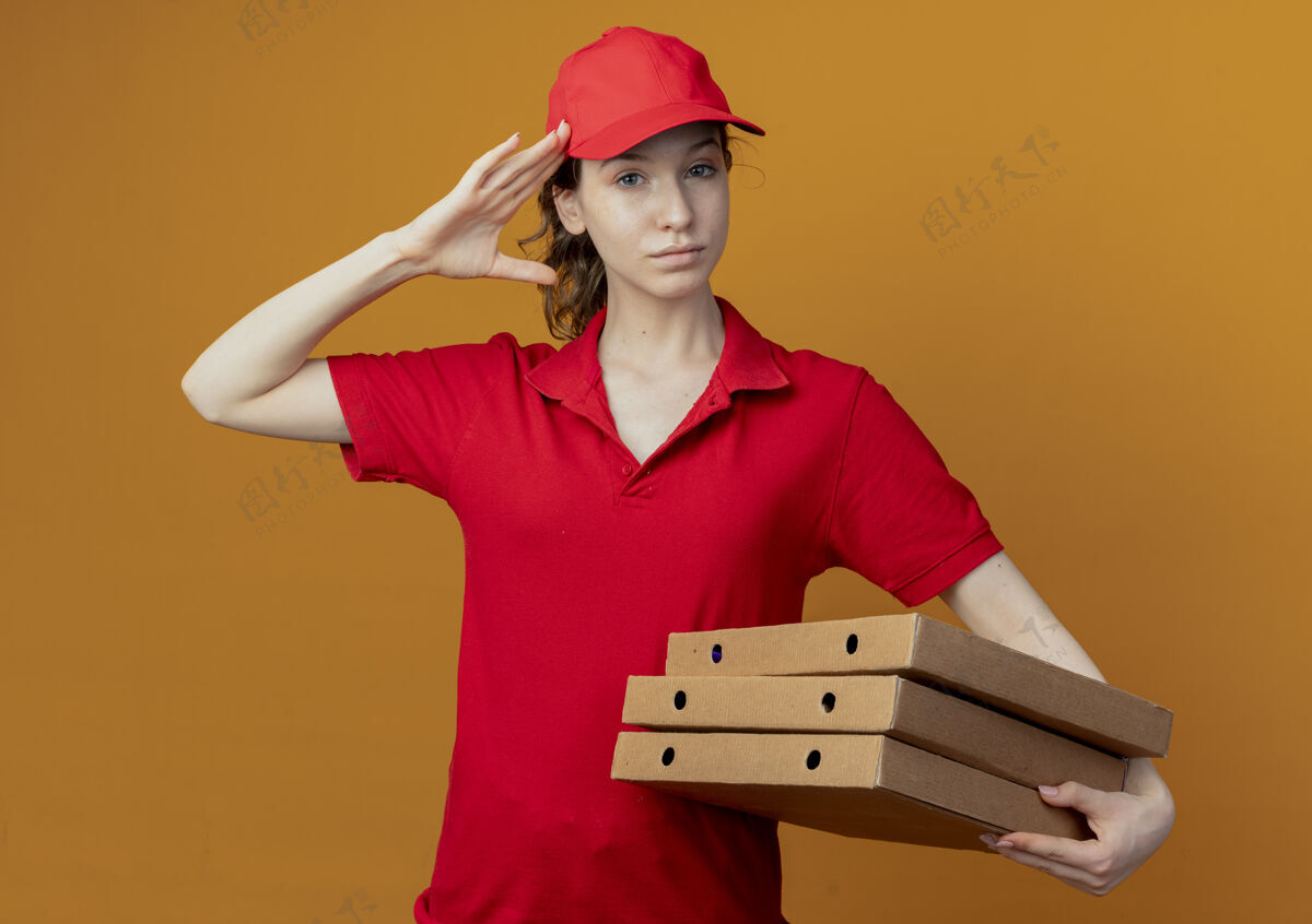 手势自信的年轻漂亮的送货女孩 穿着红色制服 戴着帽子 拿着披萨包 在橙色背景上做着敬礼手势拿着敬礼橙色