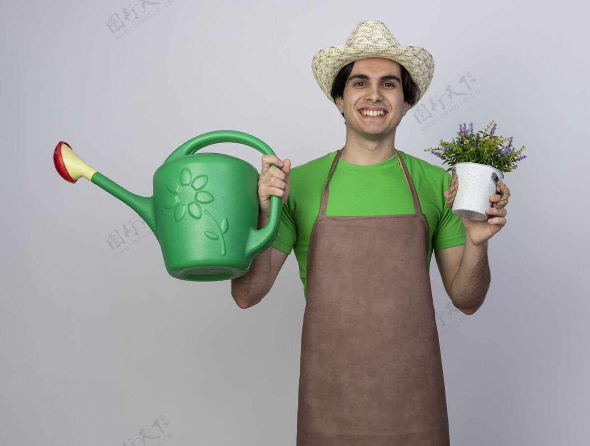 年轻人微笑着的年轻男园丁 穿着制服 戴着园艺帽 手里拿着花盆里的花和浇水罐男人戴着人