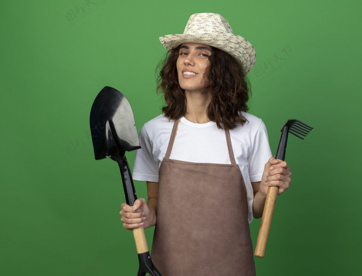 制服年轻的女园丁面带微笑 穿着制服 戴着园艺帽 手里拿着铲子 手里拿着耙子耙子帽子穿