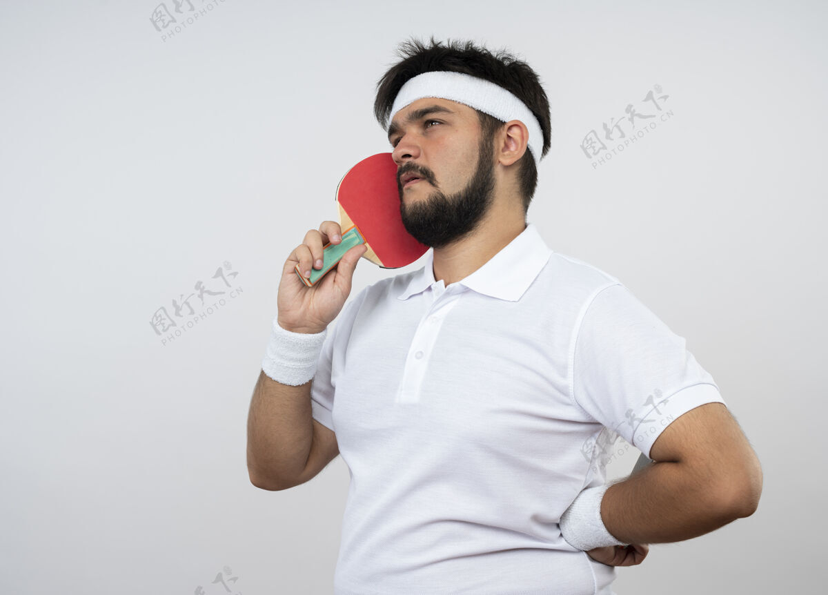 乒乓球思考年轻的运动型男子看一边戴着头带和腕带拿着乒乓球拍在脸颊上把手放在臀部隔离在白色的墙上球拍男人壁板