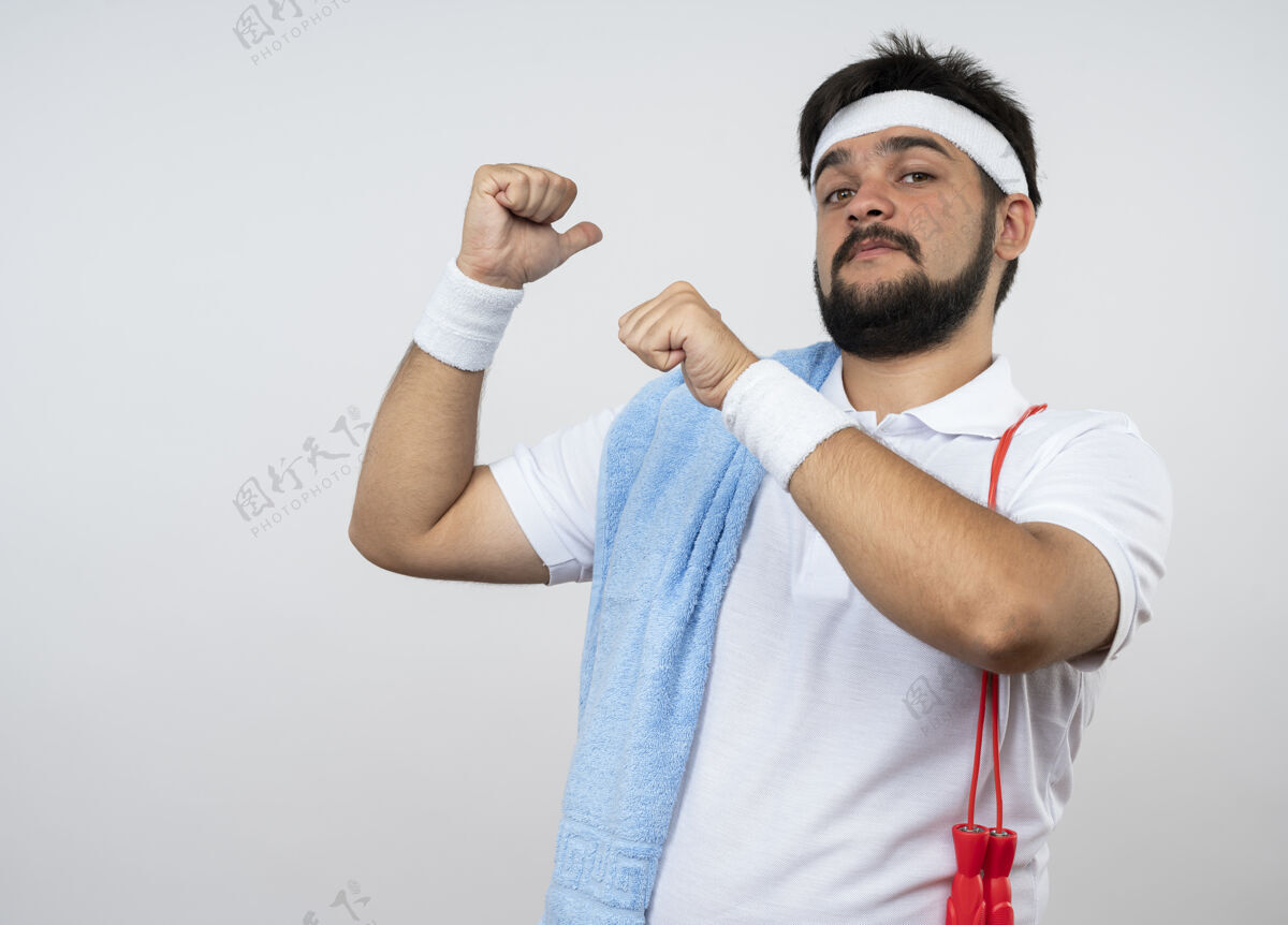 穿戴着头带和腕带的自信的年轻运动型男子用毛巾指着身后 肩上的跳绳孤立在白墙上信心绳子年轻