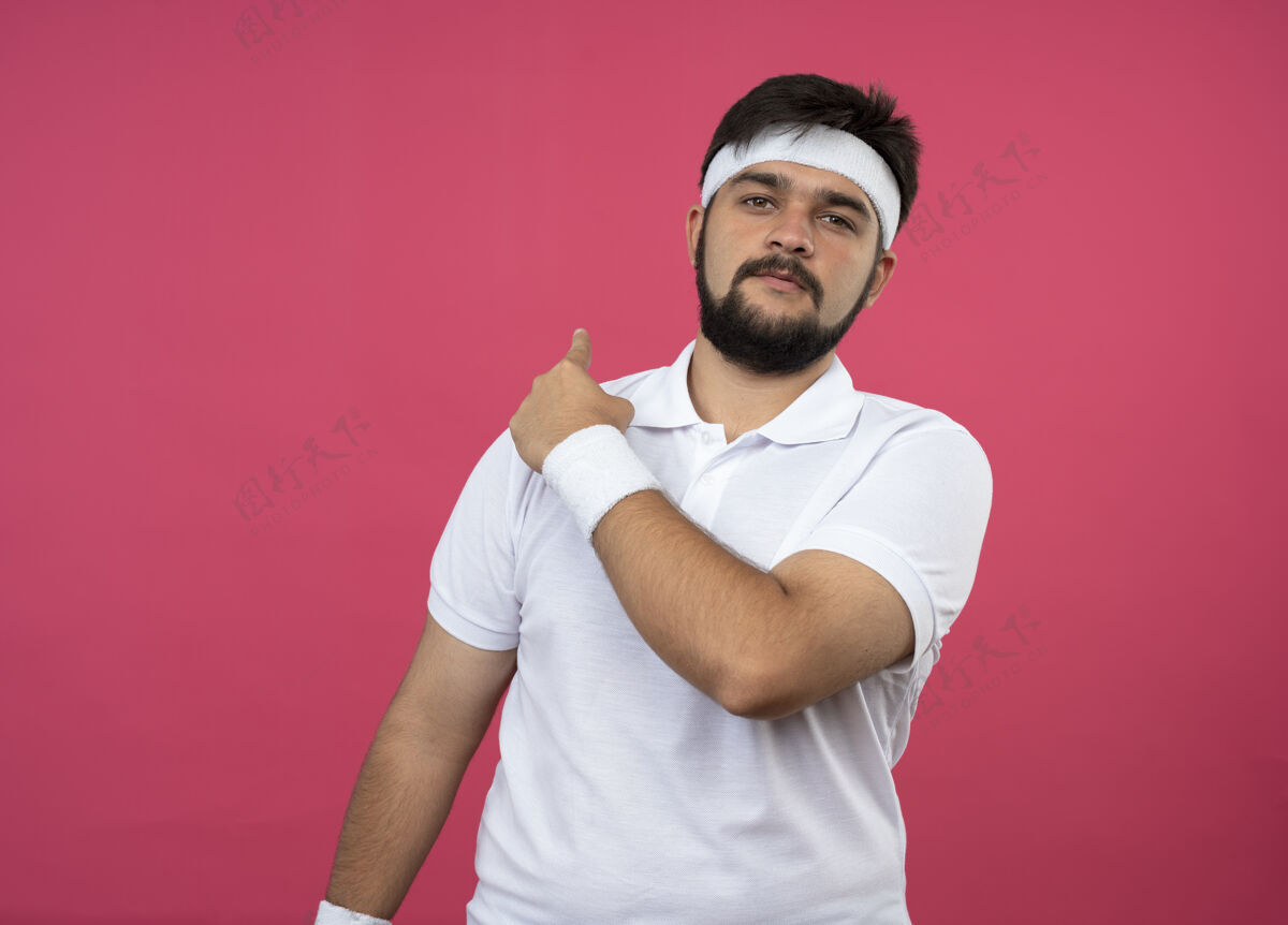 头带高兴的年轻人戴着头带和腕带点在后面孤立的粉红色墙上复制空间请戴年轻男士