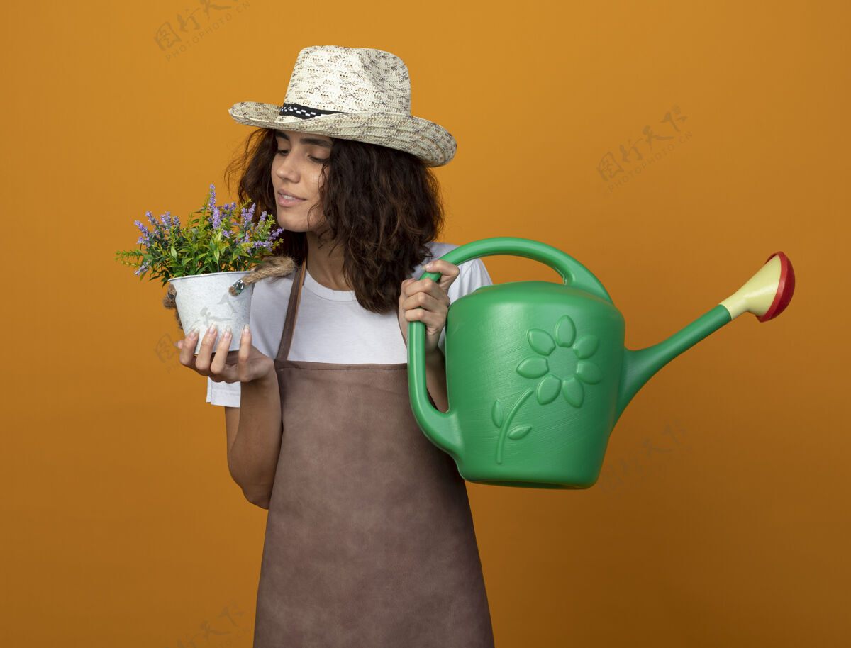 园艺高兴的年轻女园丁穿着制服戴着园艺帽拿着浇水罐隔离在橘子上制服举行女