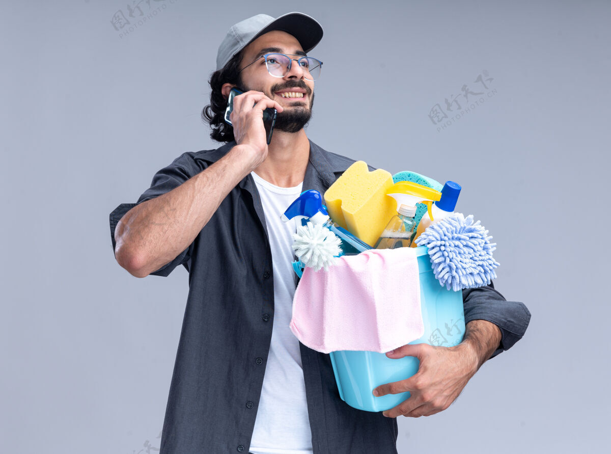 水桶面带微笑的年轻帅气的清洁工穿着t恤 戴着帽子 手里拿着一桶清洁工具 隔着白墙讲着电话年轻家伙说话