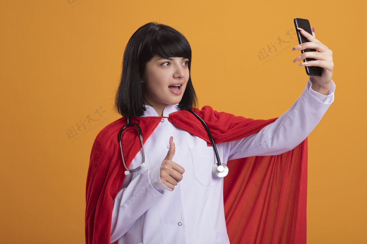 听诊器快乐的年轻超级英雄女孩戴着听诊器和医疗袍和斗篷采取自拍拇指孤立在橙色医疗穿超级英雄