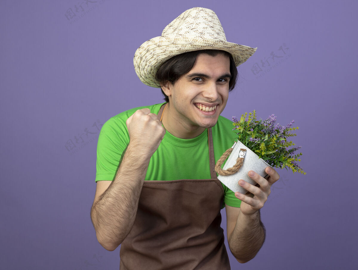 紫色微笑着的年轻男园丁 身着制服 戴着园艺帽 手里拿着花盆里的花 在紫色地上显示出“是”的手势男性手势拿着