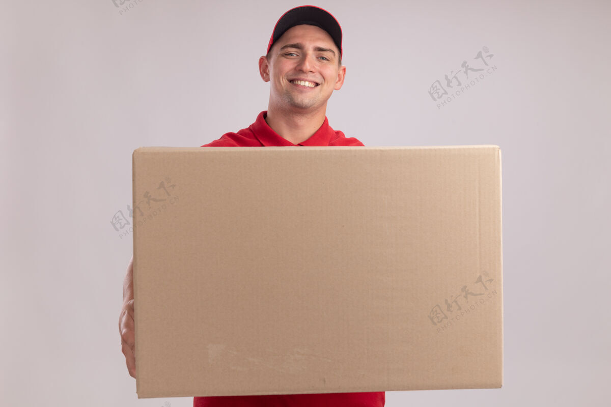 制服面带微笑的年轻送货员穿着制服 戴着帽子 手里拿着一个大箱子 隔离在白墙上送货穿拿着