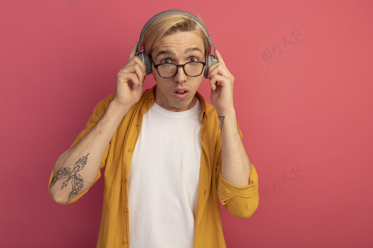 金发惊讶的目光直视前方 年轻的金发小伙子穿着黄色t恤 戴着眼镜 耳机被隔离在粉色带拷贝空间的地方穿黄色耳机