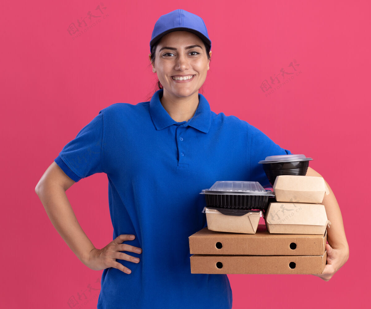 拿着带着微笑的年轻送货女孩 穿着制服 戴着帽子 拿着食物容器放在披萨盒上 手放在臀部 隔离在粉红色的墙上披萨容器帽子
