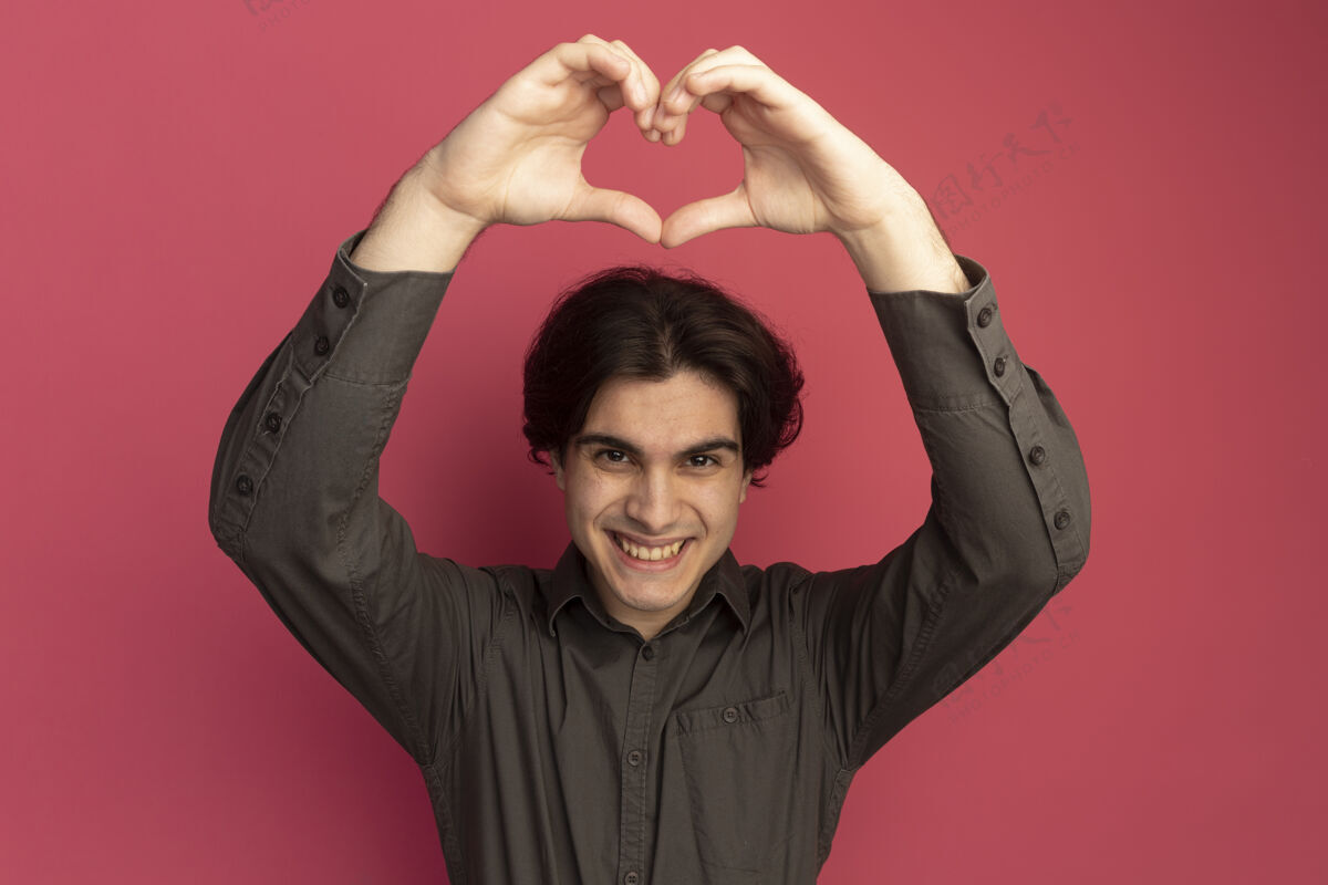心面带微笑的年轻帅哥穿着黑色t恤 在粉色的墙上展示着孤立的心形帅气微笑男人
