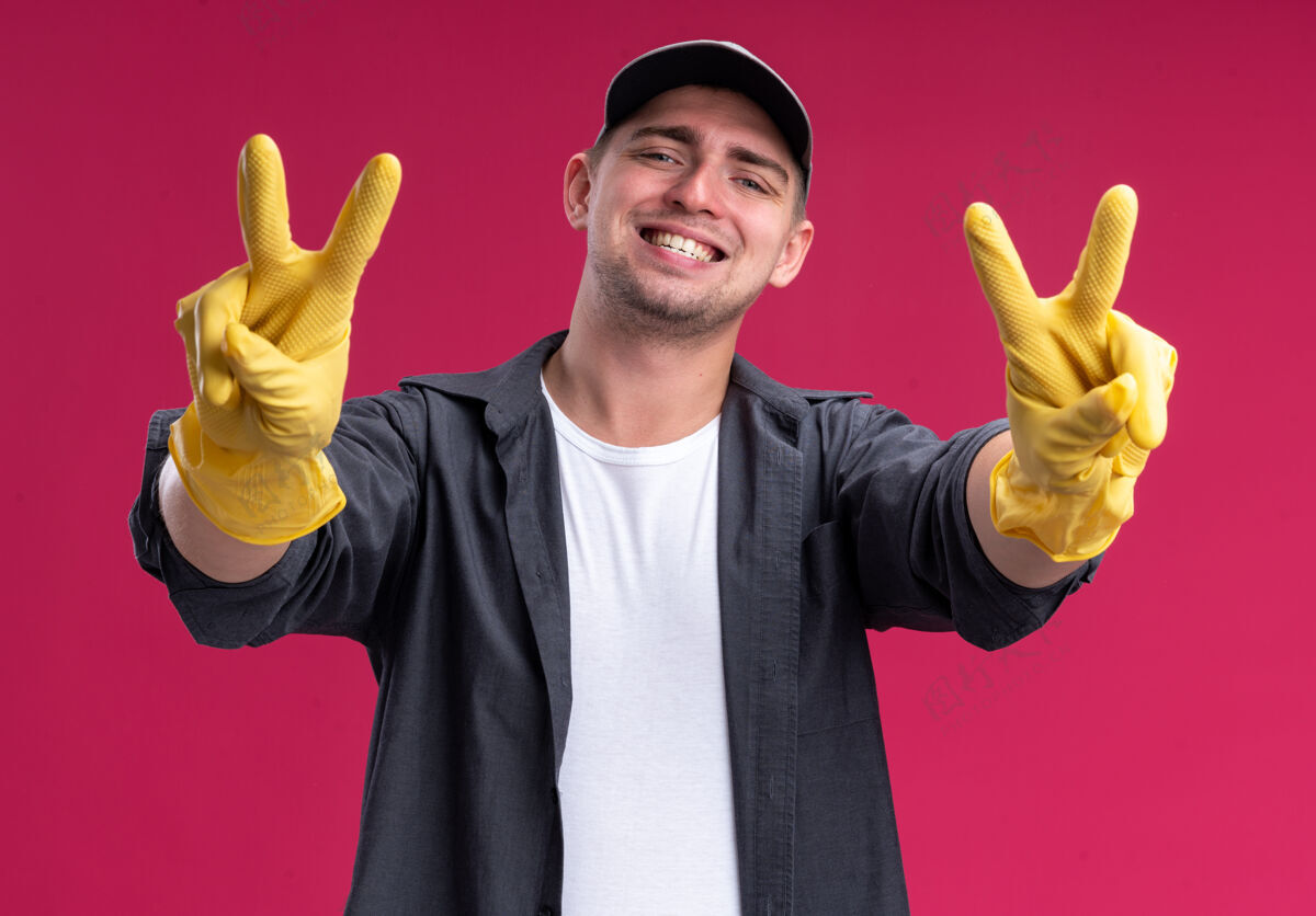 手势面带微笑的年轻汉姆森保洁员穿着t恤和帽子 戴着手套 在粉红色的墙上显示出与世隔绝的和平姿态表演帽子男人