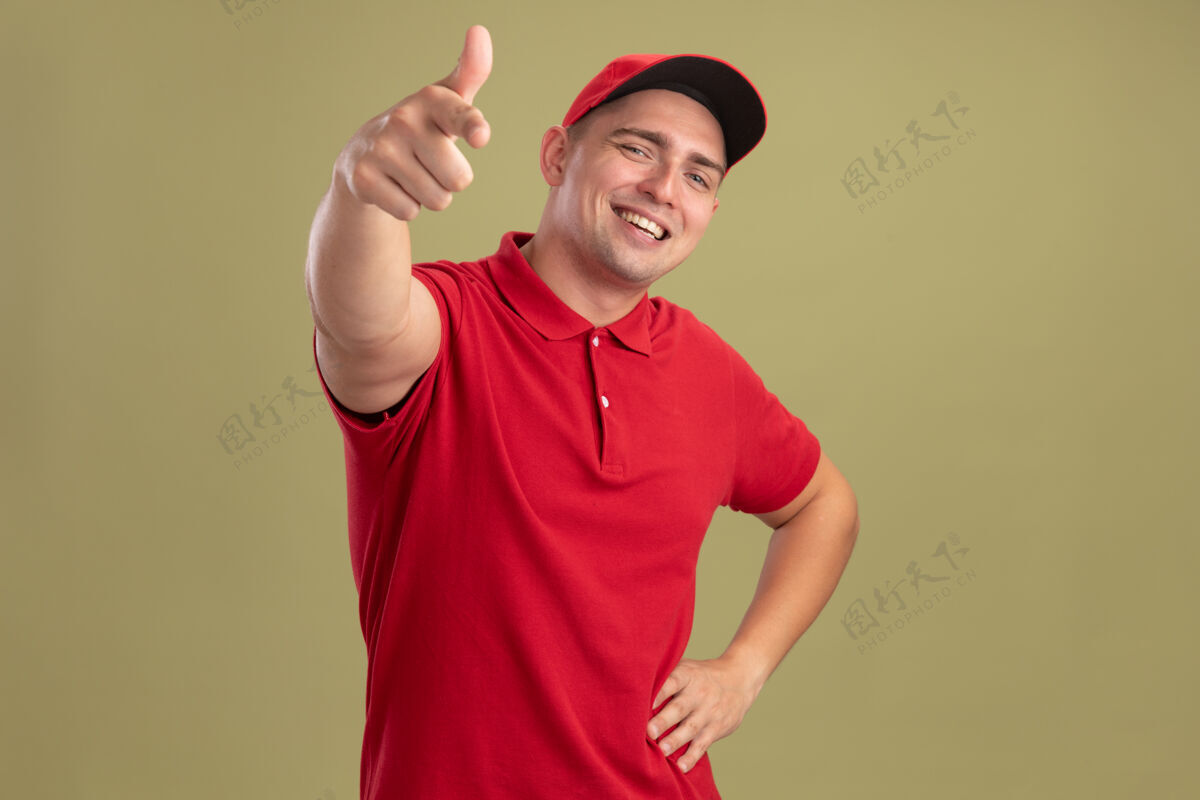 年轻微笑着的年轻送货员穿着制服 戴着帽子 向你们展示手势 手放在臀部 隔离在橄榄绿的墙上穿臀部交货