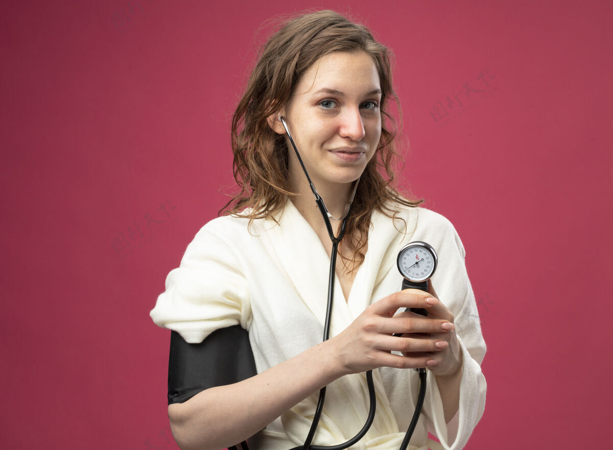 压力高兴的年轻病女孩穿着白色长袍测量自己的压力血压计隔离在粉红色血压计测量疾病